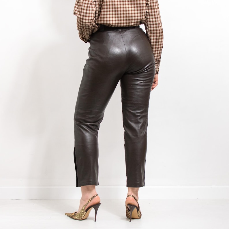 May include: Dark brown leather pants with a high waist and a slim fit. The pants have a slight sheen and a slit at the ankle. The person is wearing gold-colored high heels.