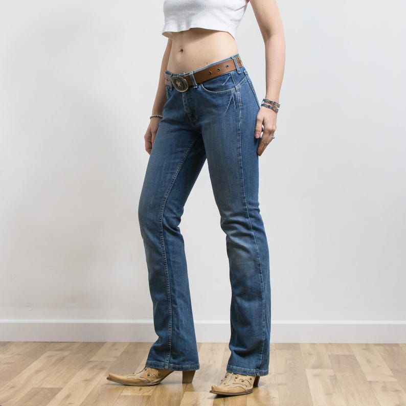 Peut inclure: Jean bleu avec une ceinture en cuir marron et une boucle d&eacute;corative. Le jean a une conception classique &agrave; cinq poches et une l&eacute;g&egrave;re jambe &eacute;vas&eacute;e. La personne porte des bottes de cowboy beiges &agrave; bout pointu.