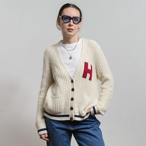 May include: Cream-coloured, chunky knit cardigan with a deep V-neck, button closure, and two front pockets. Features a large red "H" patch on the left chest and navy blue stripes on the cuffs and hem. Worn with a white top and blue jeans.
