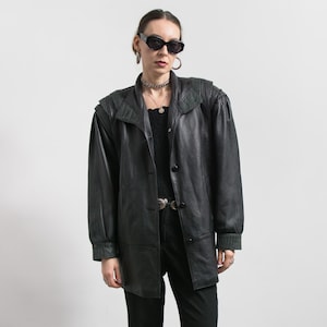 May include: Black leather jacket featuring a button-up front, shoulder pads, and a collared neckline. The jacket has long sleeves with ribbed cuffs and a tailored fit. The model is wearing black sunglasses and a silver necklace.