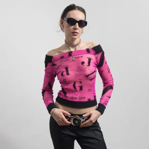 Pu&ograve; includere: Top rosa con spalle scoperte con finiture nere e maniche lunghe. La parte superiore presenta un motivo di testo nero ripetuto. La modella indossa occhiali da sole neri, una collana d'argento e una cintura nera con fibbia argentata.