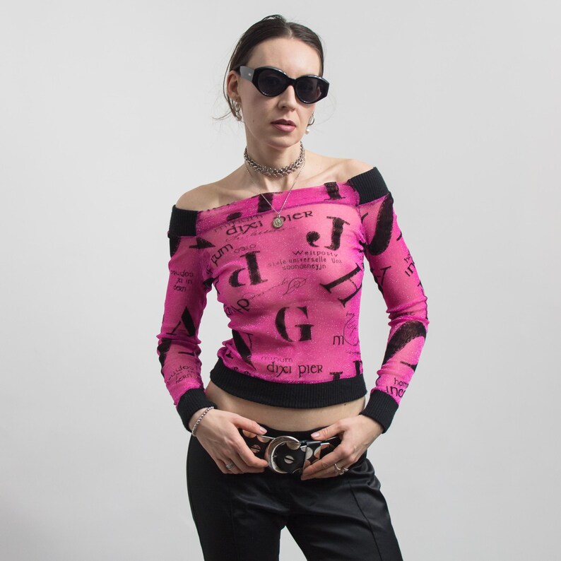 Pu&ograve; includere: Top rosa con spalle scoperte con finiture nere e maniche lunghe. La parte superiore presenta un motivo di testo nero ripetuto. La modella indossa occhiali da sole neri, una collana d'argento e una cintura nera con fibbia argentata.