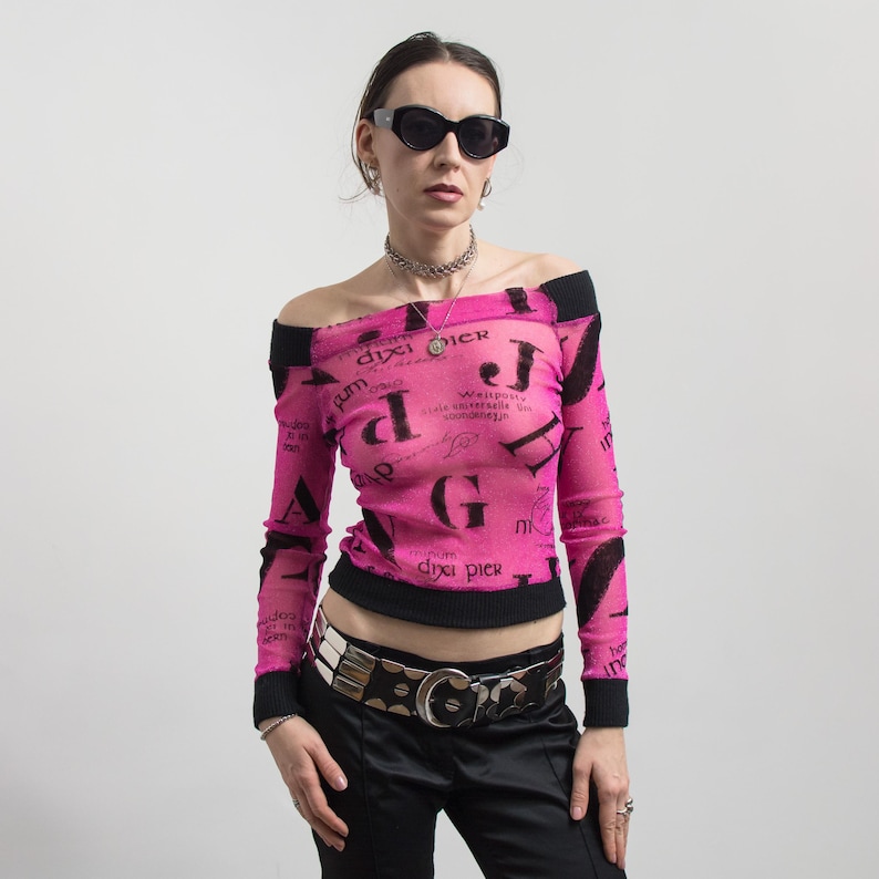 Pu&ograve; includere: Un top rosa acceso, con spalle scoperte, con finiture nere e maniche lunghe. Il top presenta un motivo di testo nero ripetuto. La modella indossa occhiali da sole neri, una collana d'argento e una cintura d'argento con pantaloni neri.