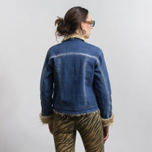 Peut inclure: Veste en jean bleue avec col et poignets en fausse fourrure beige. La veste a un design classique avec une fermeture boutonn&eacute;e et un d&eacute;lavage clair. La personne porte un pantalon &agrave; imprim&eacute; z&eacute;br&eacute;.