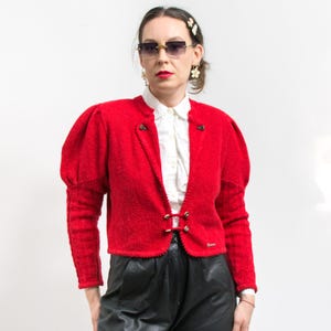 Puede incluir: Chaqueta bolero roja brillante, con tejido texturizado, mangas abullonadas y cierres dorados. Se combina con una camisa blanca con cuello y pantalones de cuero negro. Una pequeña etiqueta es visible en la parte inferior derecha.