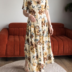 May include: A button-down, short-sleeved dress with a floral pattern in shades of brown, yellow, and green. The dress has a V-neck and a flowing, floor-length skirt. The dress is worn with sandals.