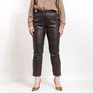 May include: Dark brown leather pants with gold button details along the outer ankles. The pants have a tailored fit and are paired with a brown plaid button-down shirt and patterned heels.