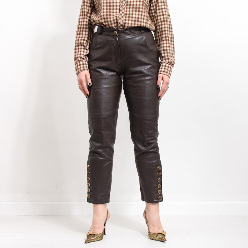 May include: Dark brown leather pants with gold button details along the outer ankles. The pants have a tailored fit and are paired with a brown plaid button-down shirt and patterned heels.