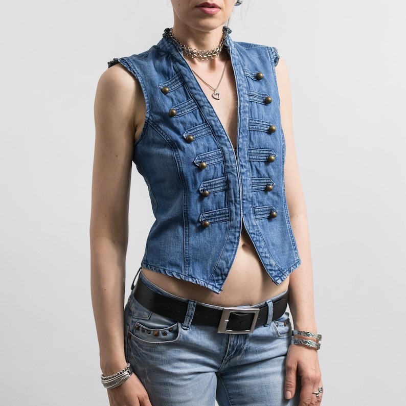 May include: A sleeveless blue denim vest featuring a military-inspired design with two vertical rows of brass-toned buttons and decorative cord detailing. The vest has a high collar, a fitted silhouette, and is worn over light blue denim jeans.