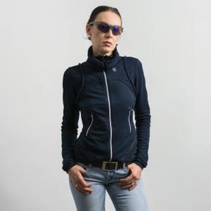 May include: A dark navy blue zip-up athletic jacket with a high collar, long sleeves, and two front pockets featuring white zipper trim. The jacket includes a small white circular logo on the upper chest and is paired with light blue denim jeans.