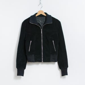 Puede incluir: Chaqueta bomber negra con cremallera, dos bolsillos laterales y pu&ntilde;os y dobladillo acanalados. La chaqueta tiene un cuello azul oscuro y est&aacute; colgada de una percha de madera sobre un fondo blanco.