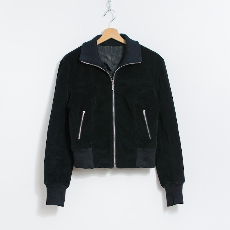 Puede incluir: Chaqueta bomber negra con cremallera, dos bolsillos laterales y pu&ntilde;os y dobladillo acanalados. La chaqueta tiene un cuello azul oscuro y est&aacute; colgada de una percha de madera sobre un fondo blanco.