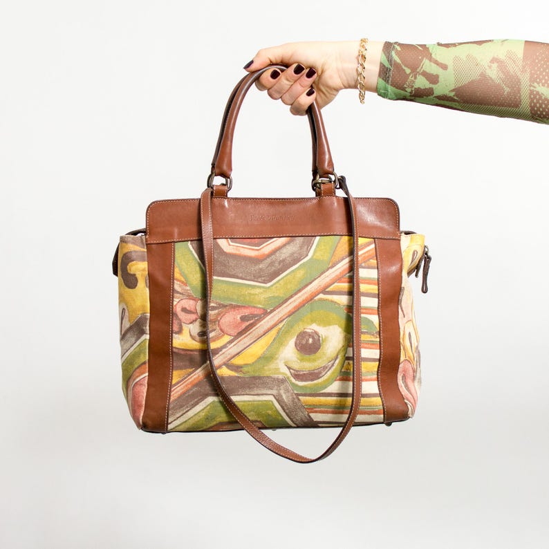 May include: A brown leather tote bag with a colorful abstract design. The bag has brown leather handles and trim, a shoulder strap, and a zipper closure. The abstract design features yellow, green, brown, and pink shapes.
