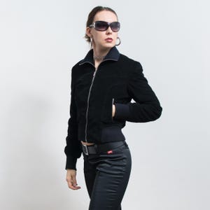 Puede incluir: Chaqueta bomber de pana negra con cremallera plateada, usada con pantalones negros y cintur&oacute;n negro. La chaqueta tiene cuello y pu&ntilde;os acanalados. La persona lleva gafas de sol y un collar plateado.