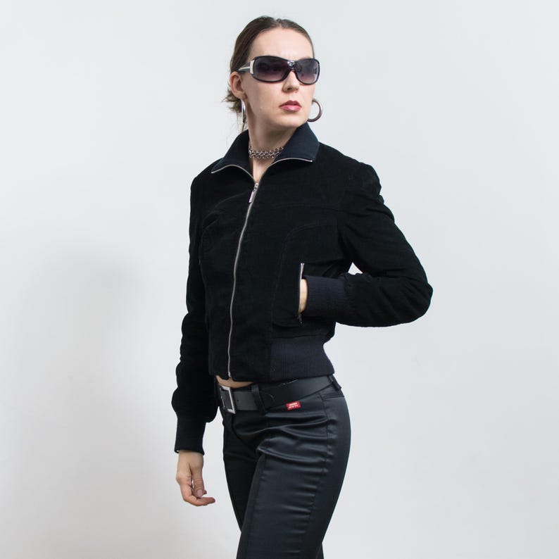 Puede incluir: Chaqueta bomber de pana negra con cremallera plateada, usada con pantalones negros y cintur&oacute;n negro. La chaqueta tiene cuello y pu&ntilde;os acanalados. La persona lleva gafas de sol y un collar plateado.