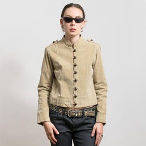 Może przedstawiać: Beżowa kurtka zamszowa z kołnierzem typu stójka i zapięciem na guziki. Kurtka ma długie rękawy i ozdobne guziki na ramionach. Osoba nosi okulary przeciwsłoneczne i pasek.