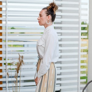 May include: A woman wearing a white button-down shirt with a long, beige and brown striped skirt. She has her hair pulled back in a bun and is wearing large hoop earrings.
