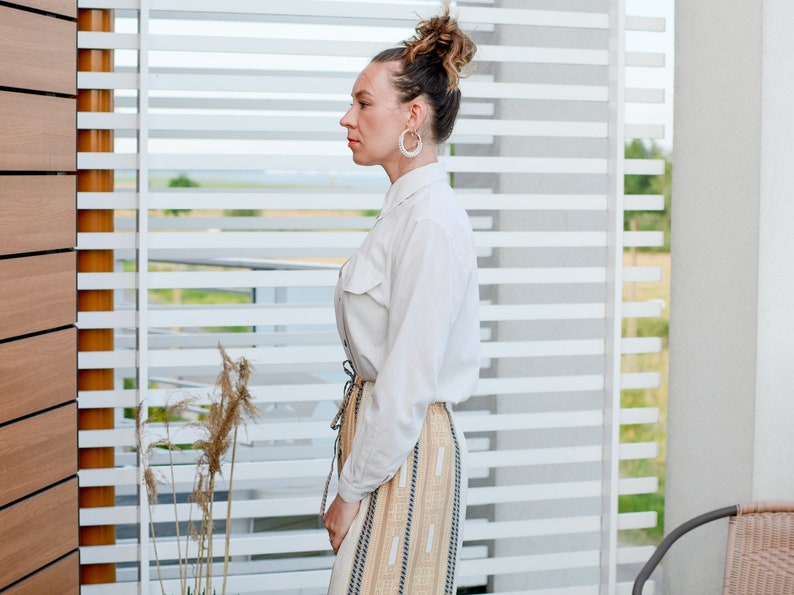 May include: A woman wearing a white button-down shirt with a long, beige and brown striped skirt. She has her hair pulled back in a bun and is wearing large hoop earrings.