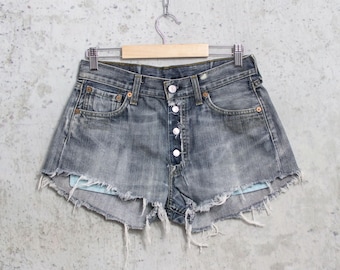 May include: Distressed denim shorts with a button fly and frayed edges. The shorts are a faded blue-gray color and hang from a wooden hanger. The shorts have front and back pockets and a button closure.