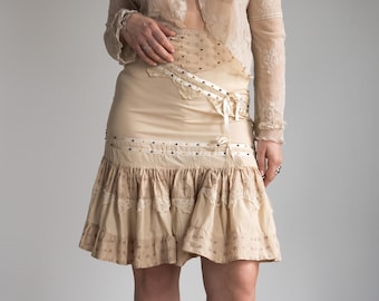 May include: A beige skirt with a layered, ruffled hem and decorative trim. The skirt features a fitted waist with a ribbon tie and is paired with a sheer, cream-colored top. The outfit has a vintage, bohemian aesthetic.