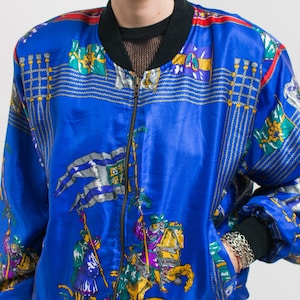 May include: A blue satin bomber jacket with a zipper closure. The jacket features a colorful print of flags, knights, and horses.
