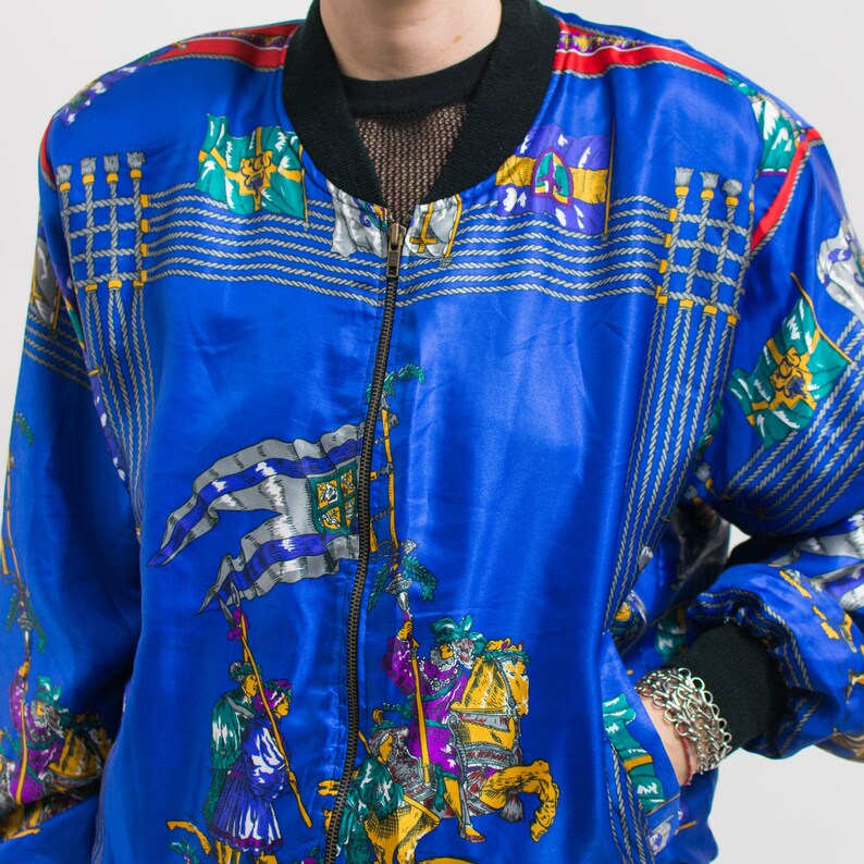 May include: A blue satin bomber jacket with a zipper closure. The jacket features a colorful print of flags, knights, and horses.