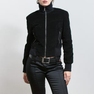 Puede incluir: Chaqueta negra de terciopelo con cuello alto y cremallera plateada. La chaqueta tiene dos bolsillos laterales con cremallera y un dobladillo y pu&ntilde;os de canal&eacute;. La modelo lleva pantalones de sat&eacute;n negros y un cintur&oacute;n negro.