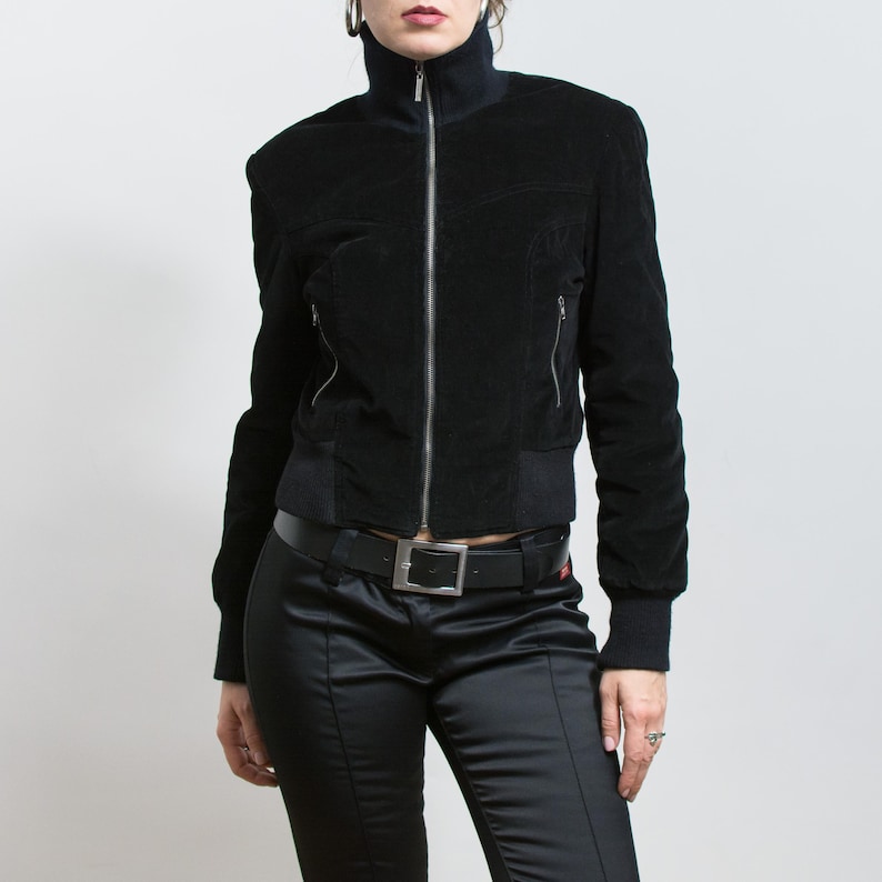 Puede incluir: Chaqueta negra de terciopelo con cuello alto y cremallera plateada. La chaqueta tiene dos bolsillos laterales con cremallera y un dobladillo y pu&ntilde;os de canal&eacute;. La modelo lleva pantalones de sat&eacute;n negros y un cintur&oacute;n negro.