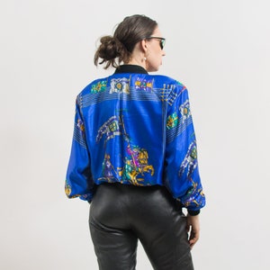 May include: A blue satin bomber jacket with a colorful print of knights and flags. The jacket has a zipper closure and long sleeves.