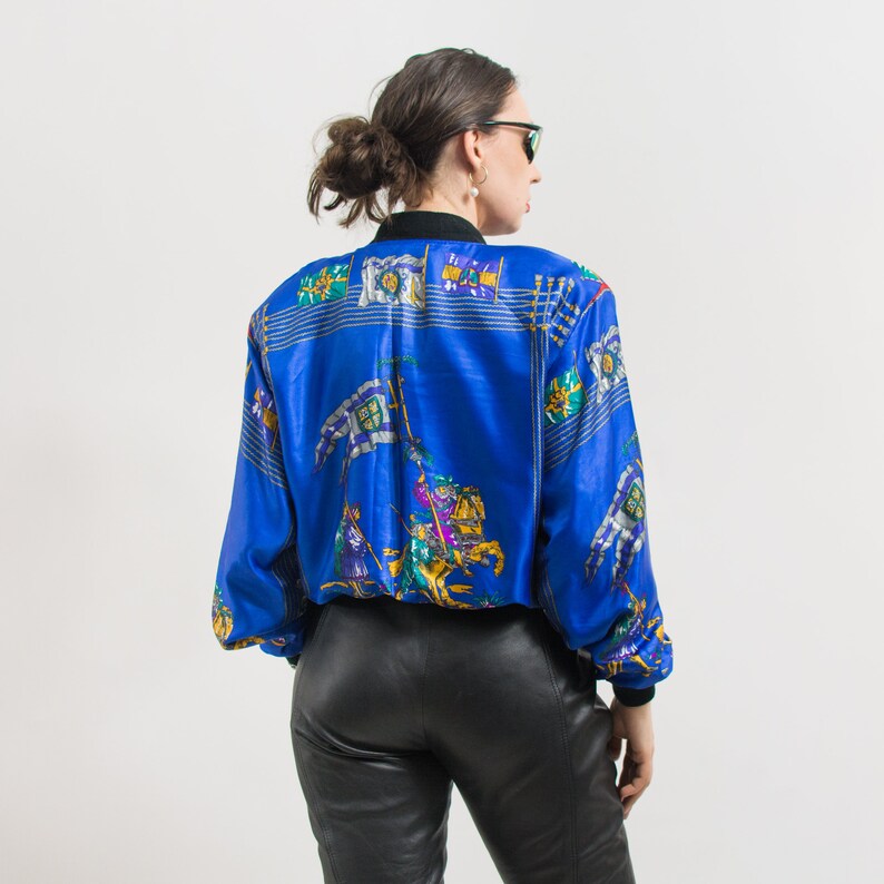 May include: A blue satin bomber jacket with a colorful print of knights and flags. The jacket has a zipper closure and long sleeves.