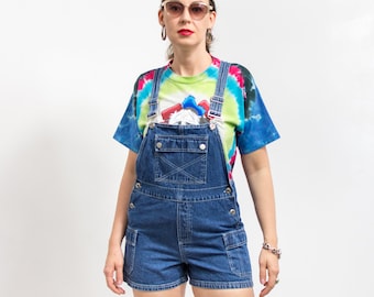 Puede incluir: Una mujer con una falda vaquera corta de peto azul, una camiseta de tie-dye y gafas de sol.