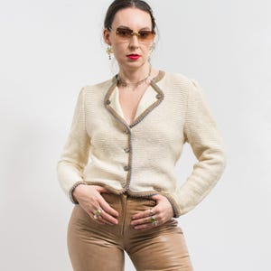 May include: Cream-colored, textured jacket with a V-neckline, featuring brown trim and blue stitching. The jacket has long sleeves and is buttoned up the front. The model is wearing brown leather pants and sunglasses.