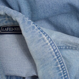 May include: Close-up of a light blue denim jacket. The jacket features a label that reads "LAFEI-NIE". The fabric is folded, revealing the stitching and texture of the denim material.