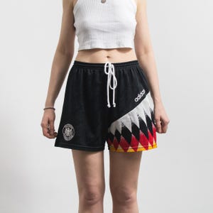 Könnte beinhalten: Schwarze Adidas-Shorts mit weißem Kordelzug. Die Shorts haben ein geometrisches Design in Rot, Weiß und Gelb am rechten Bein und ein rundes Emblem am linken Bein. Das Adidas-Logo ist am rechten Bein sichtbar.