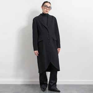 May include: A long, black coat with a notched collar and three buttons. The coat has two front pockets and a split back. The person in the image is wearing a black turtleneck and black trousers. The coat is a classic style.