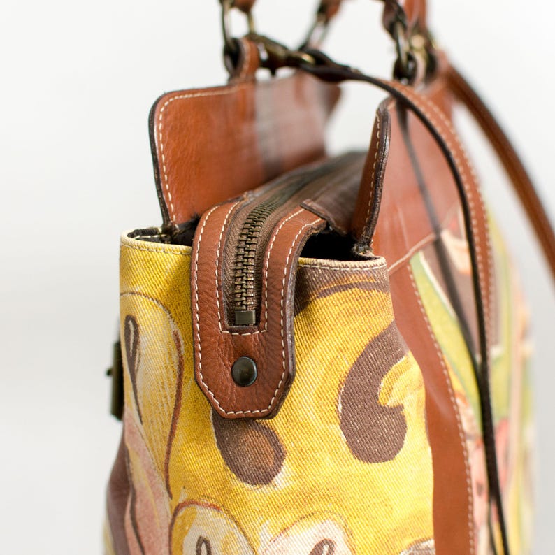 May include: Close-up of a handbag with a yellow and brown patterned fabric. The bag features brown leather accents, a zipper closure, and a brown leather handle. White stitching details the leather trim.