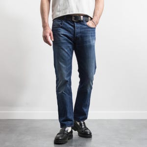 May include: A pair of dark blue denim jeans with a faded wash, featuring a classic five-pocket design, a button fly, and a black leather belt with a silver-toned metal buckle. The jeans are paired with a plain white t-shirt and black leather loafers.