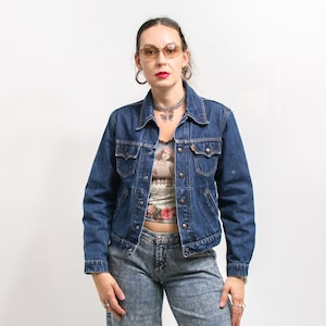 Op de afbeelding: Een vrouw draagt een blauw denim jack over een tanktop met bloemenprint en een zilveren ketting. Ze draagt een blauwe jeans en heeft een bril met bruine monturen.