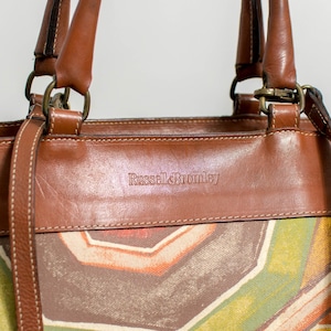 May include: A brown leather handbag with a colorful geometric pattern. The bag features brown leather handles and trim, with the brand name "Russell & Bromley" embossed on the front. The bag has a shoulder strap and metal hardware.