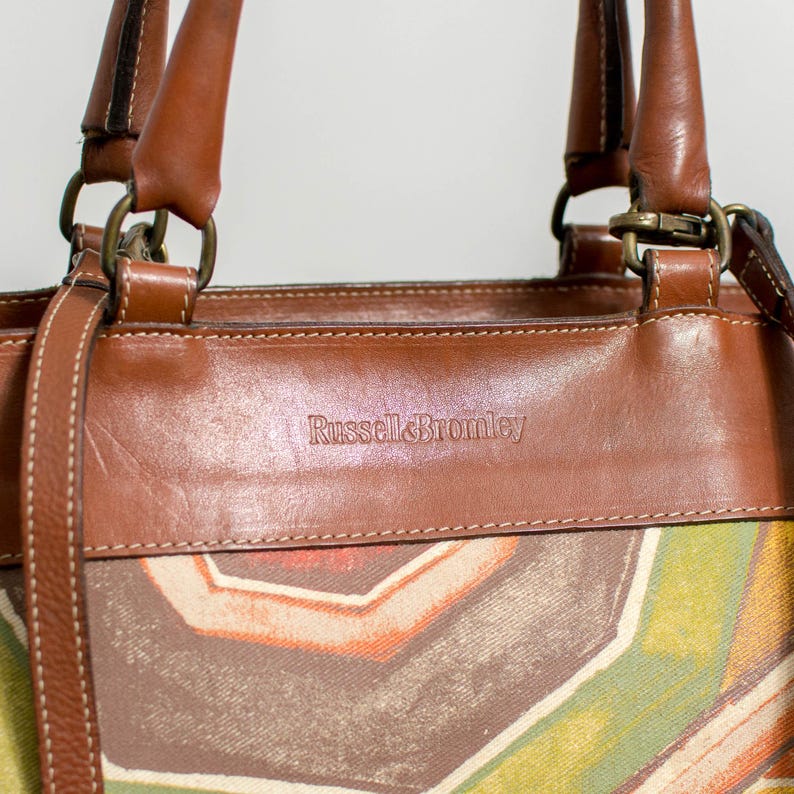 May include: A brown leather handbag with a colorful geometric pattern. The bag features brown leather handles and trim, with the brand name "Russell & Bromley" embossed on the front. The bag has a shoulder strap and metal hardware.