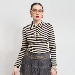 Pode incluir: Uma camisa de mangas compridas listrada com gola e zíper dourado. A camisa apresenta listras horizontais pretas e creme. A pessoa está usando uma saia cinza com cinto decorativo e joias douradas.