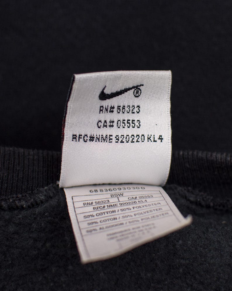 May include: Close-up of a black garment tag with the Nike logo and text. The tag includes RN and CA numbers, along with fabric composition details. The tag is attached to a dark fabric.