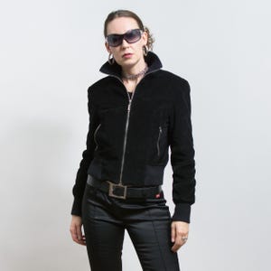 Puede incluir: Chaqueta bomber de pana negra con cremallera, bolsillos laterales y dobladillo y pu&ntilde;os de canal&eacute;. La chaqueta se combina con pantalones negros y un cintur&oacute;n negro con hebilla plateada. La persona lleva gafas de sol y joyas de plata.