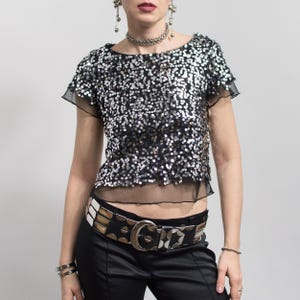 Può includere: Top nero con paillettes con maniche corte e orlo trasparente. Il top è abbinato a una cintura larga borchiata e pantaloni neri. Gli accessori includono una collana d'argento, orecchini e bracciali.