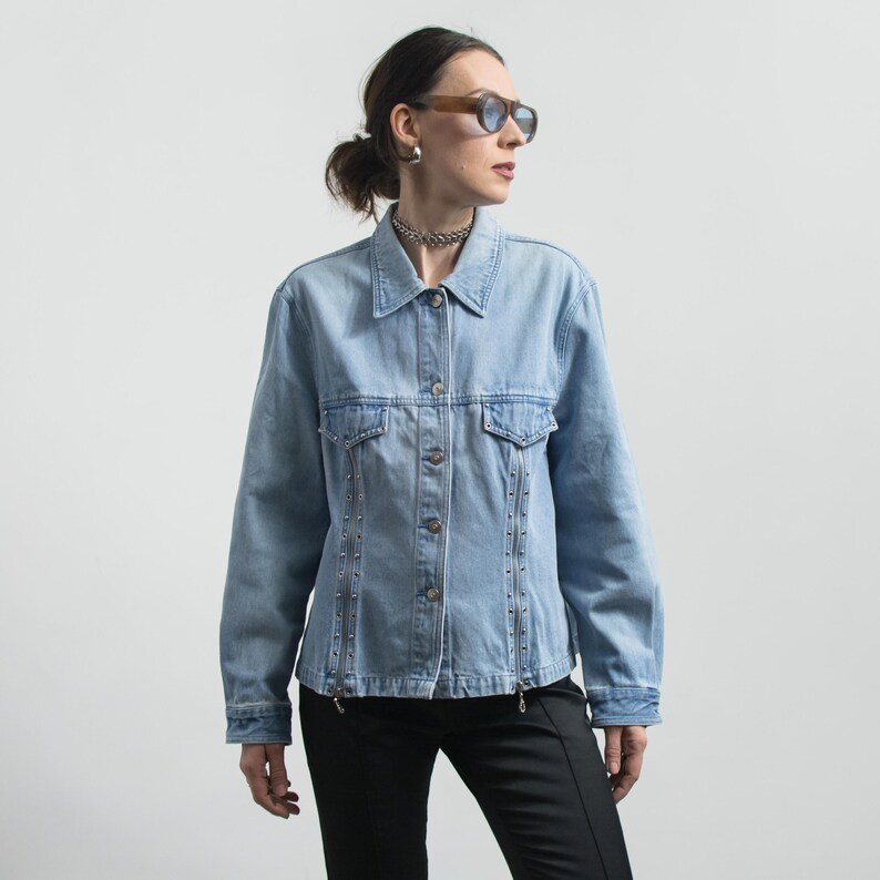 May include: Light blue denim jacket with silver-toned stud accents along the front and sleeves. The jacket has a collared neckline, two chest pockets, and a button-down closure. The model is wearing black pants and sunglasses.