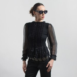 Puede incluir: Blusa negra con mangas largas transparentes, corpi&ntilde;o con volantes y detalle decorativo en la cintura. La blusa tiene un cuello alto y se combina con pantalones negros. La persona lleva gafas de sol oscuras y pendientes.
