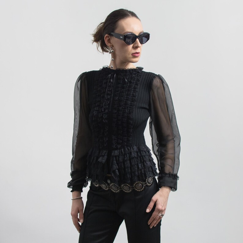 Puede incluir: Blusa negra con mangas largas transparentes, corpi&ntilde;o con volantes y detalle decorativo en la cintura. La blusa tiene un cuello alto y se combina con pantalones negros. La persona lleva gafas de sol oscuras y pendientes.