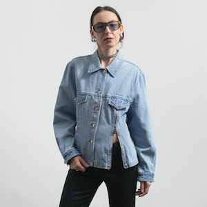 May include: Light blue denim jacket with silver-tone stud accents along the zipper and seams. The jacket has a classic collar, button closures, and two chest pockets. The model is wearing black pants and sunglasses.
