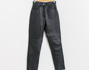 May include: Black leather pants hanging on a wooden hanger. The pants have a high waist, pockets, and a button closure. The leather appears slightly textured, and the pants are straight-legged.