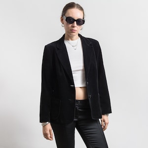 Puede incluir: Una americana de terciopelo negro con solapa de muesca, dos botones frontales y dos bolsillos de parche. La chaqueta se lleva sobre un top corto de canalé blanco y pantalones negros, junto con gafas de sol ovaladas y collares de plata.